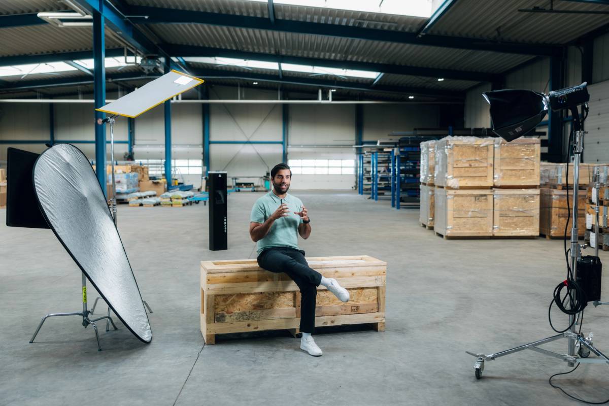 A man sits on a wooden crate in a warehouse, surrounded by studio lights, reflectors and production equipment.