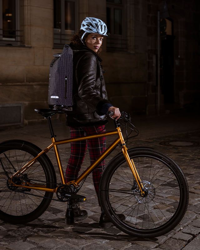 Woman with a helmet and backpack standing with a gold-coloured bike on cobblestones at night