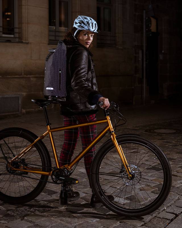 Woman with a helmet and backpack standing with a gold-coloured bike on cobblestones at night