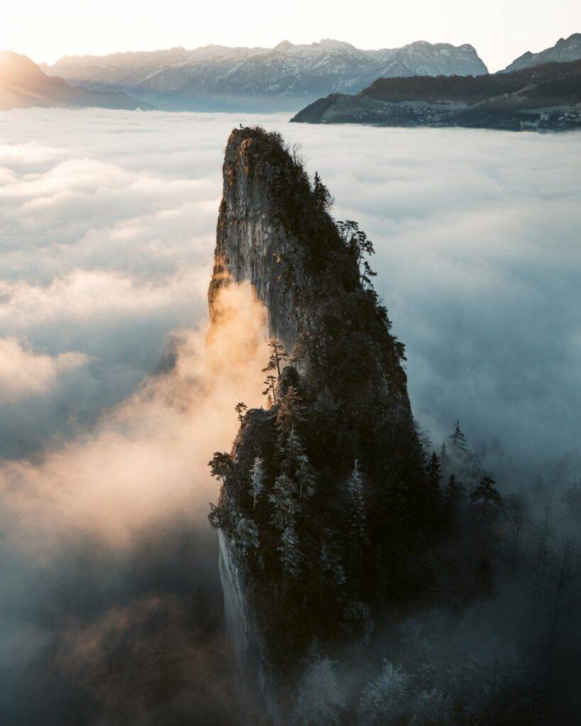 A steep, tree-covered rock spire rises through glowing clouds, with rugged mountains in the distance.