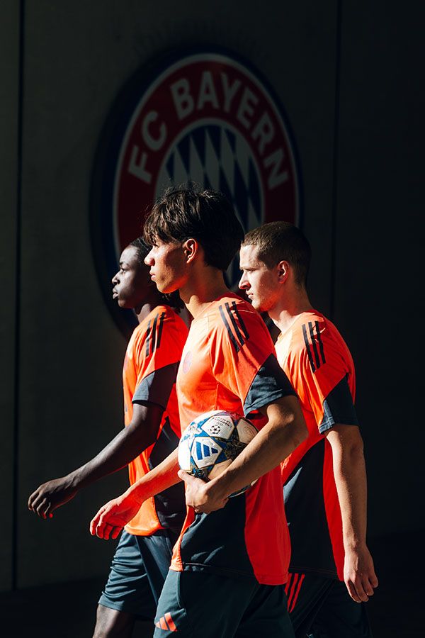 Drei Spieler gehen durch Licht und Schatten an einem FC-Bayern-Wappen vorbei, einer trägt einen Fußball