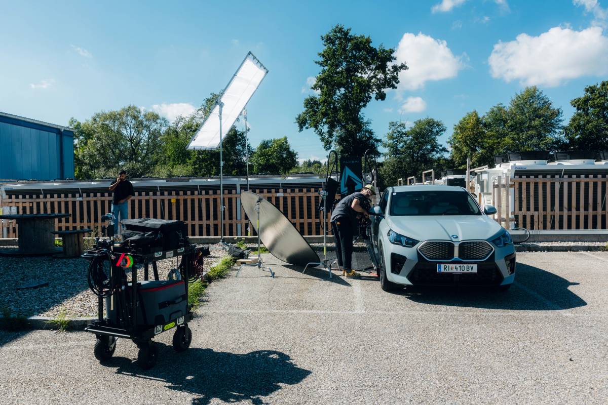 Crew members film a white car outdoors with large reflectors, lighting panels and equipment carts.