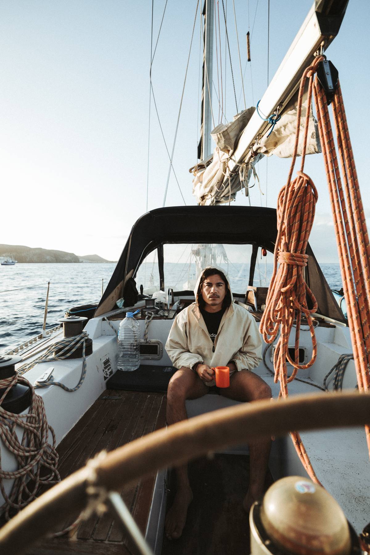 Person with a mug sitting in the cockpit of a sailing boat, steering wheel and coiled ropes in the foreground
