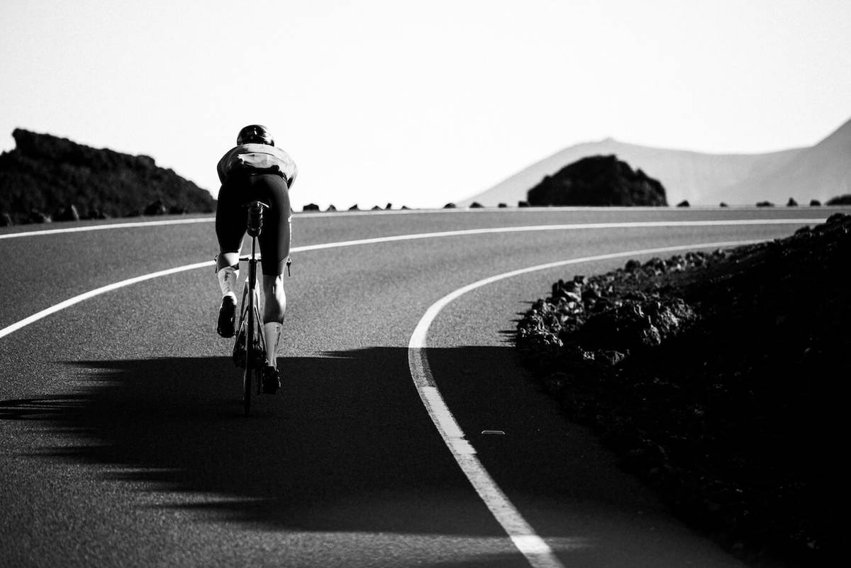 Black-and-white photo of the cyclist from behind, riding into a bend between lava fields