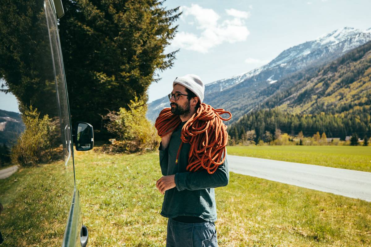 Person in a beanie with a climbing rope over the shoulder standing next to a van in a meadow