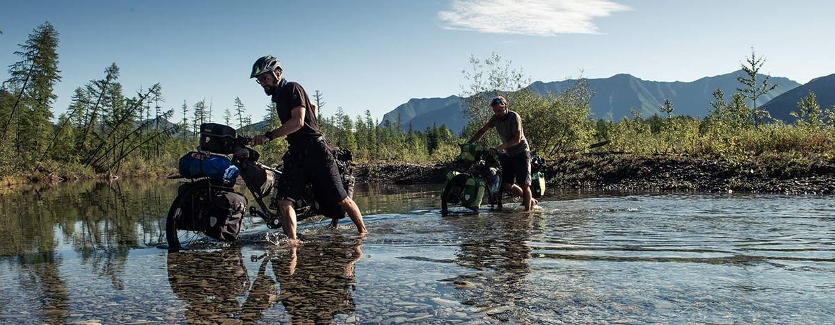 Two cyclists pushing loaded bikes through shallow water