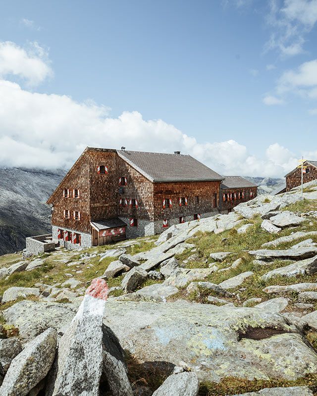 Shingle-clad mountain hut on rocky ground, a red-and-white trail marker in the foreground
