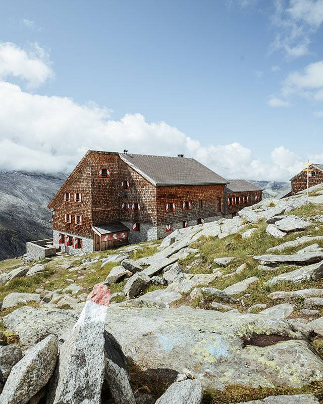 Shingle-clad mountain hut on rocky ground, a red-and-white trail marker in the foreground