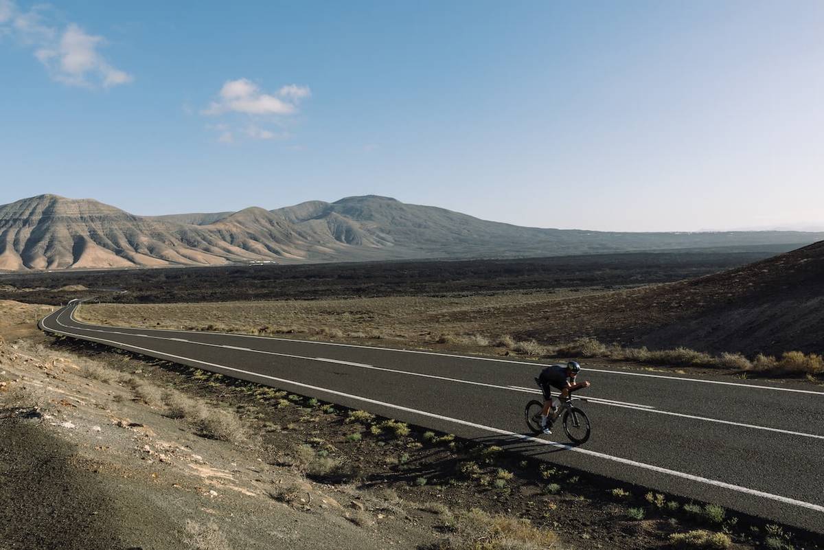 Wide shot of the cyclist on a winding road, a mountain range behind him