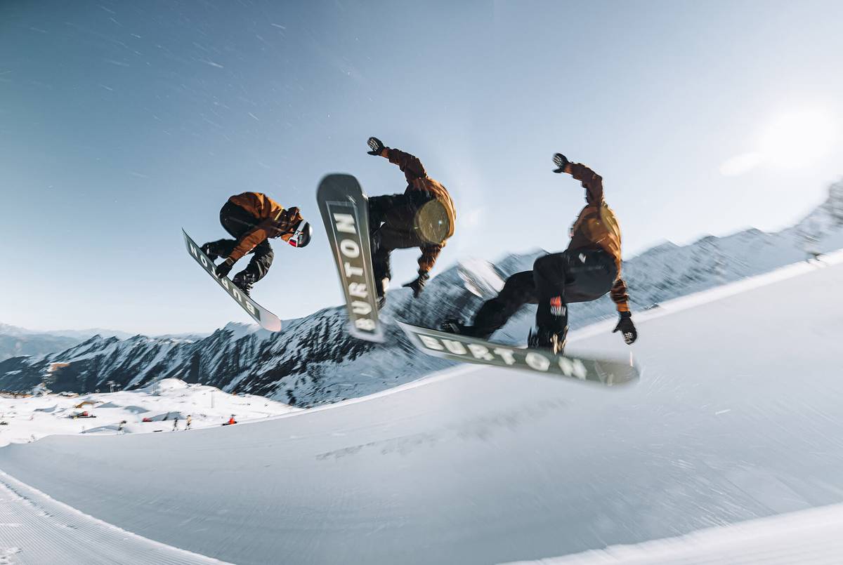 Drei Snowboarder springen gleichzeitig aus einer Halfpipe, im Hintergrund verschneite Berge