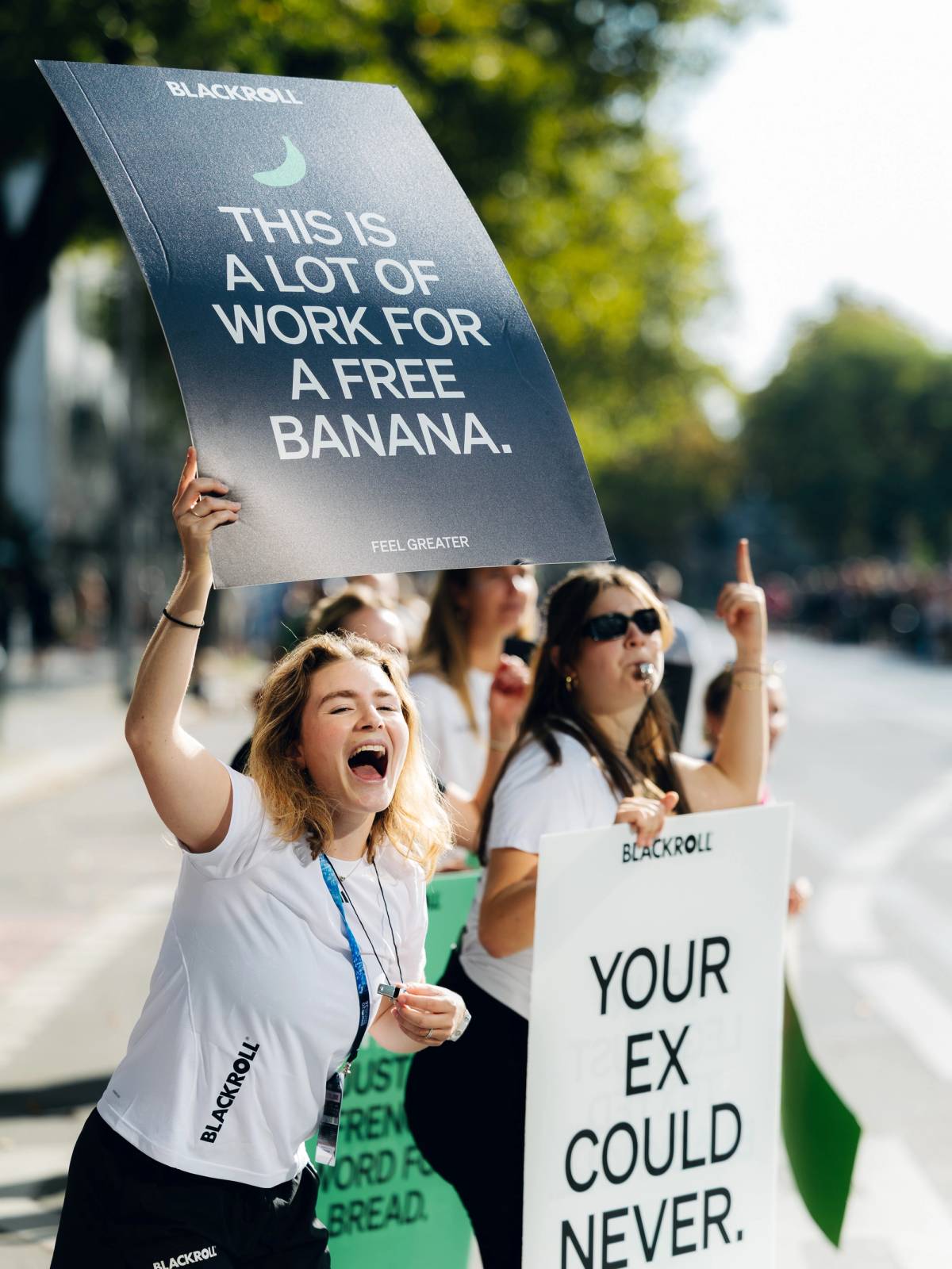 Cheering production crew members hold humorous signs and blow whistles on a sunny street.