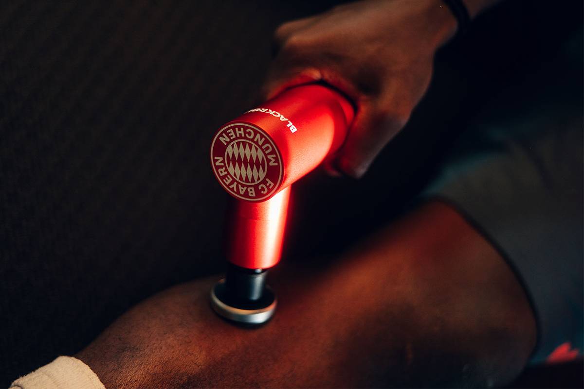 Nahaufnahme einer roten Massagepistole mit FC-Bayern-Wappen am Oberschenkel eines Spielers