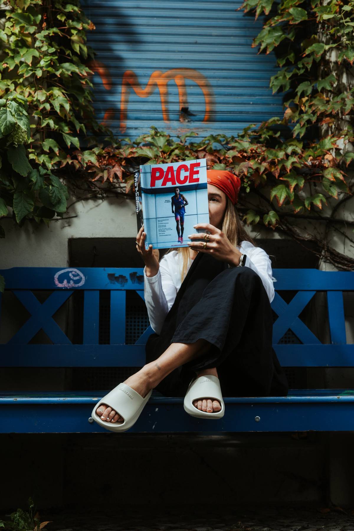 A woman in a red headscarf sits on a blue bench, holding a magazine over her face amid leafy vines and a graffiti-covered shutter.