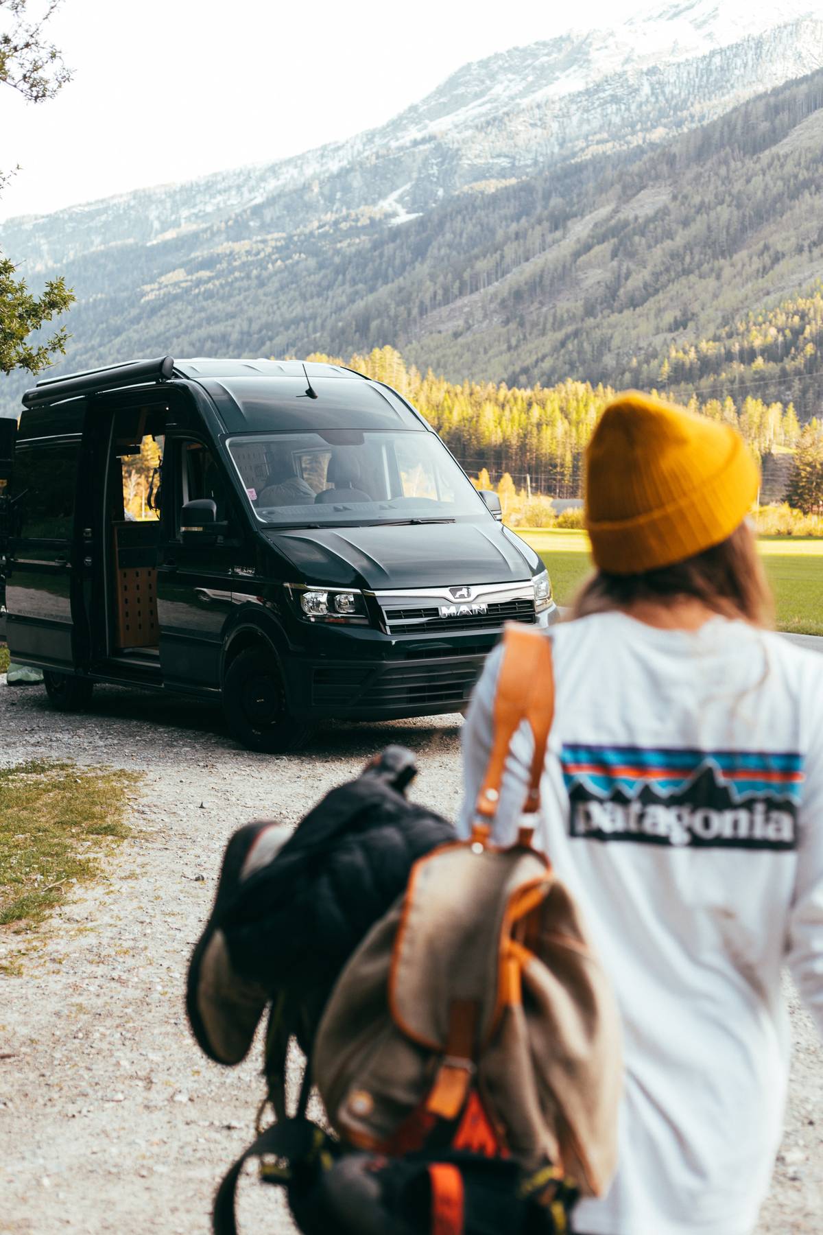 Person in a yellow beanie carrying a backpack walks towards a black van, snowy mountains behind