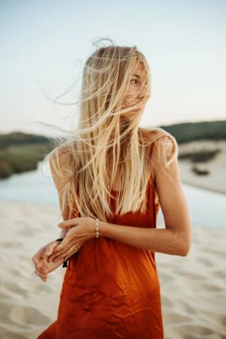 Blonde Frau in orangefarbenem Kleid steht im Wind an einem sandigen Ufer.