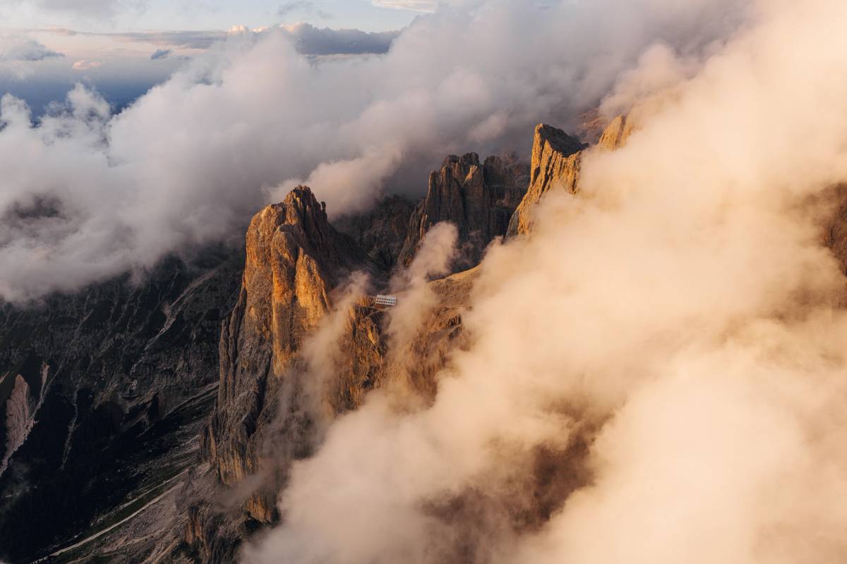 Sunlit rocky mountain peaks rise through thick clouds, with a small building visible on a ridge.