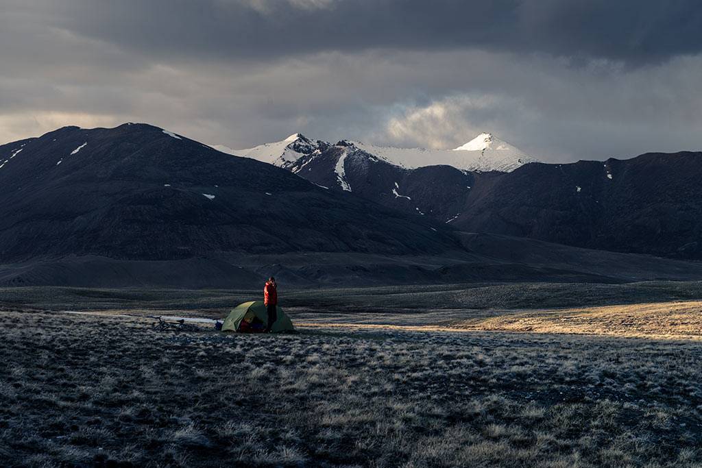 Zelt und Person in roter Jacke auf einer weiten Hochebene, dahinter schneebedeckte Berge