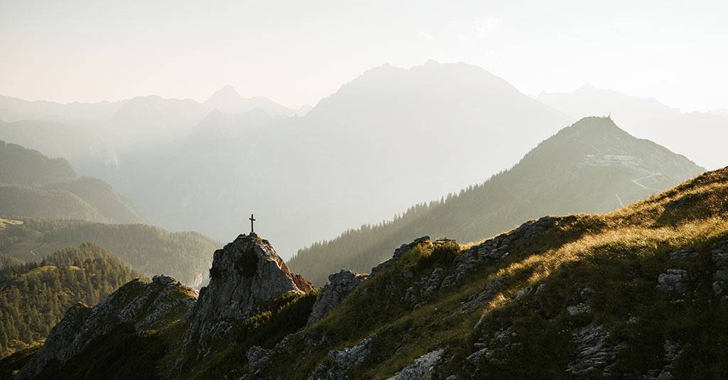 Summit cross on a rocky outcrop, layers of mountains in the haze behind it