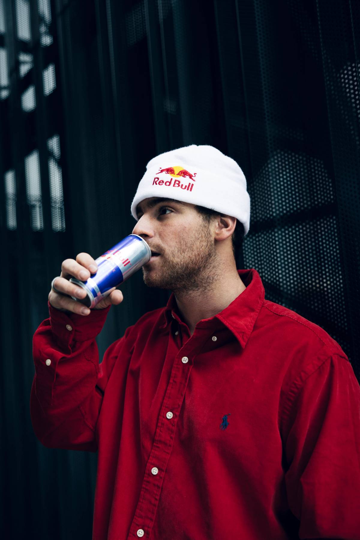 A man in a white beanie and red shirt drinks from a can in front of a dark perforated wall.