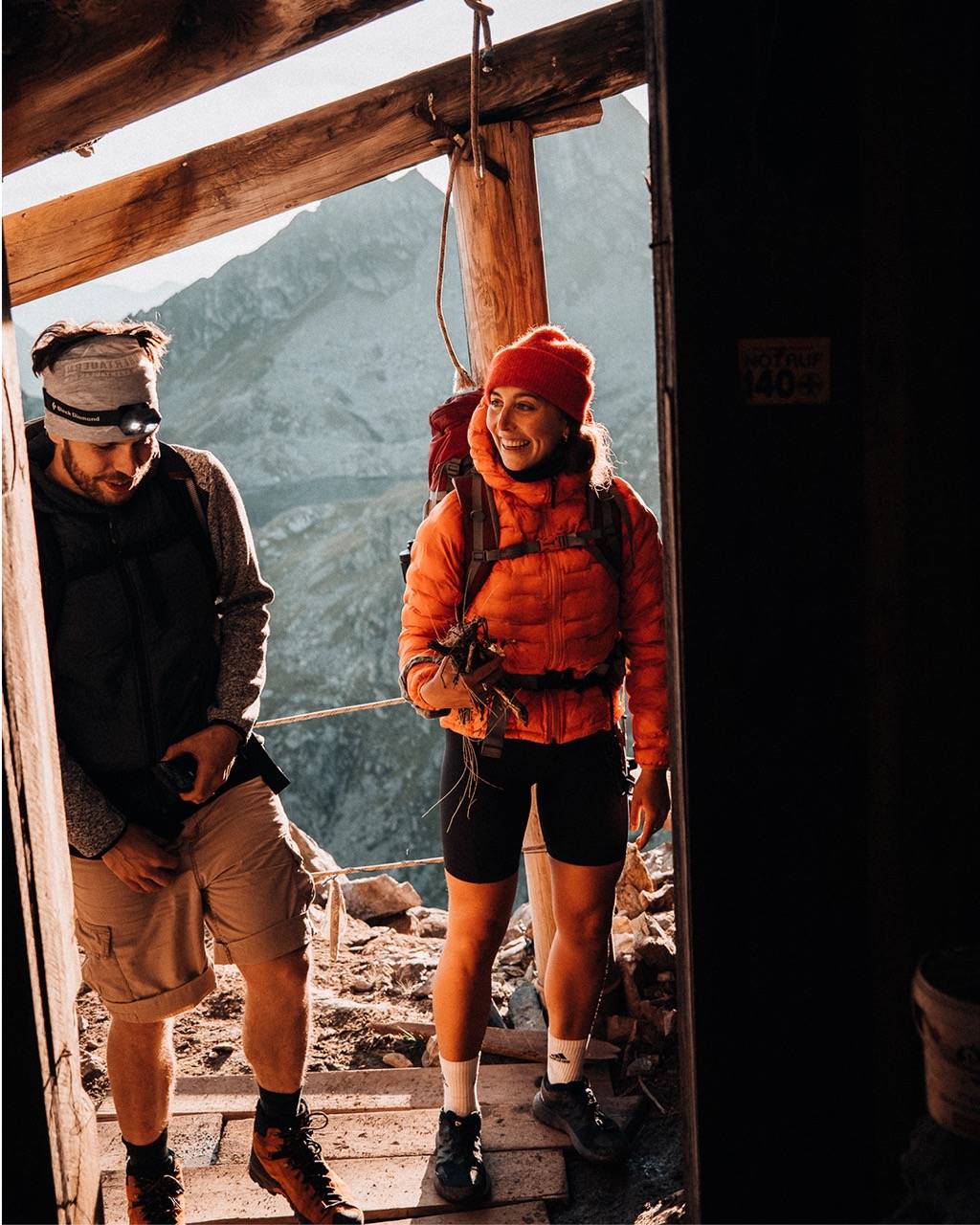 Two mountaineers stepping out of a wooden hut, laughing, a rock face behind them