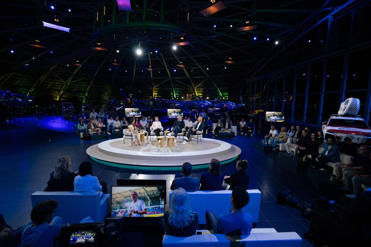 Guests sit on a circular studio set surrounded by an audience, screens, production equipment, aircraft and a display car.