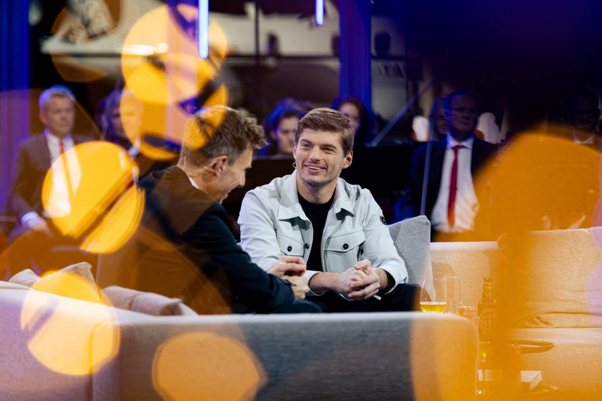 Two men talk on a studio sofa before an audience, framed by blurred orange lights.