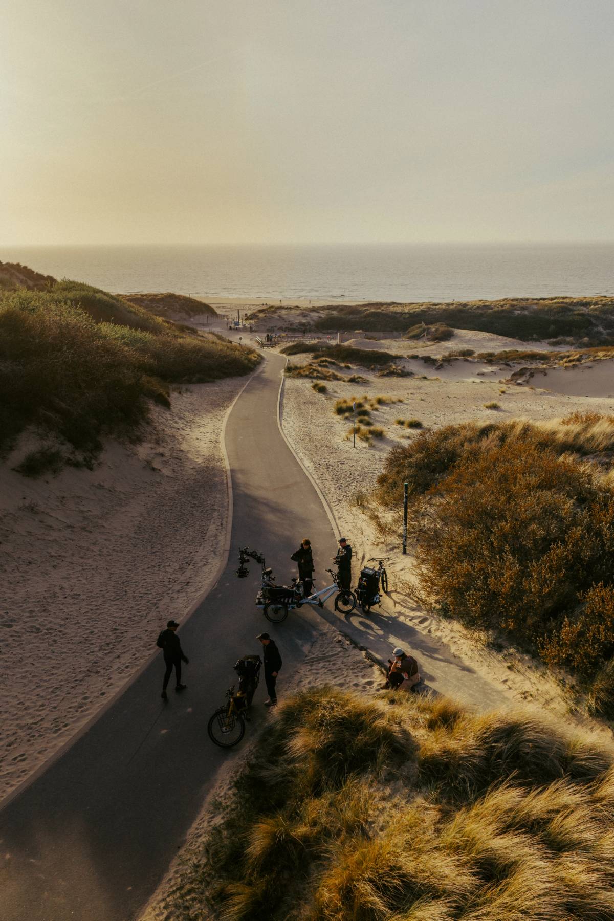 A production crew stands with camera-equipped bicycles on a winding path through coastal dunes.