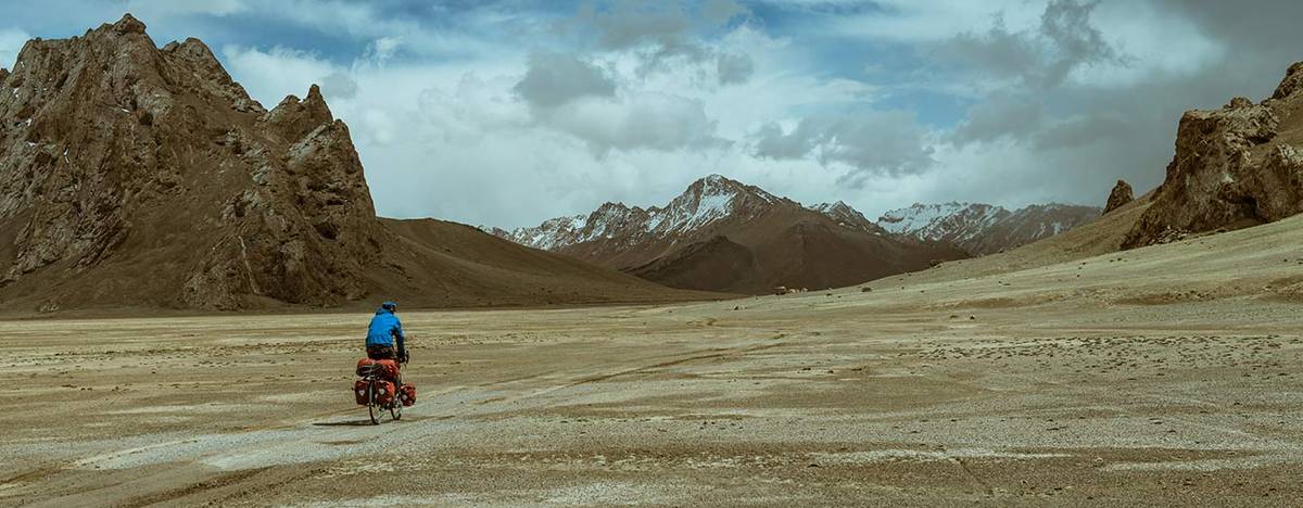 Lone cyclist with a loaded bike on a vast gravel plain below rugged mountains