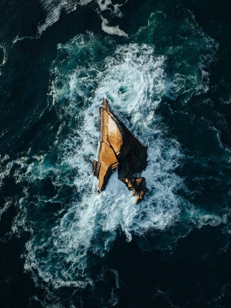 Aerial view of white waves swirling around a jagged golden-brown rock in dark blue ocean water.