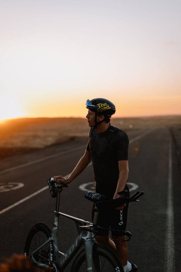 Triathlete standing with his time-trial bike on an empty road at sunset