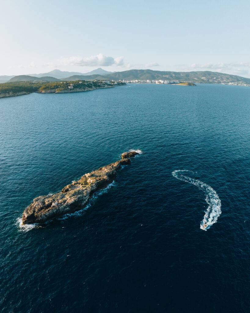 Aerial view of a boat curving around a rocky islet in blue water, with a mountainous coast in the distance.