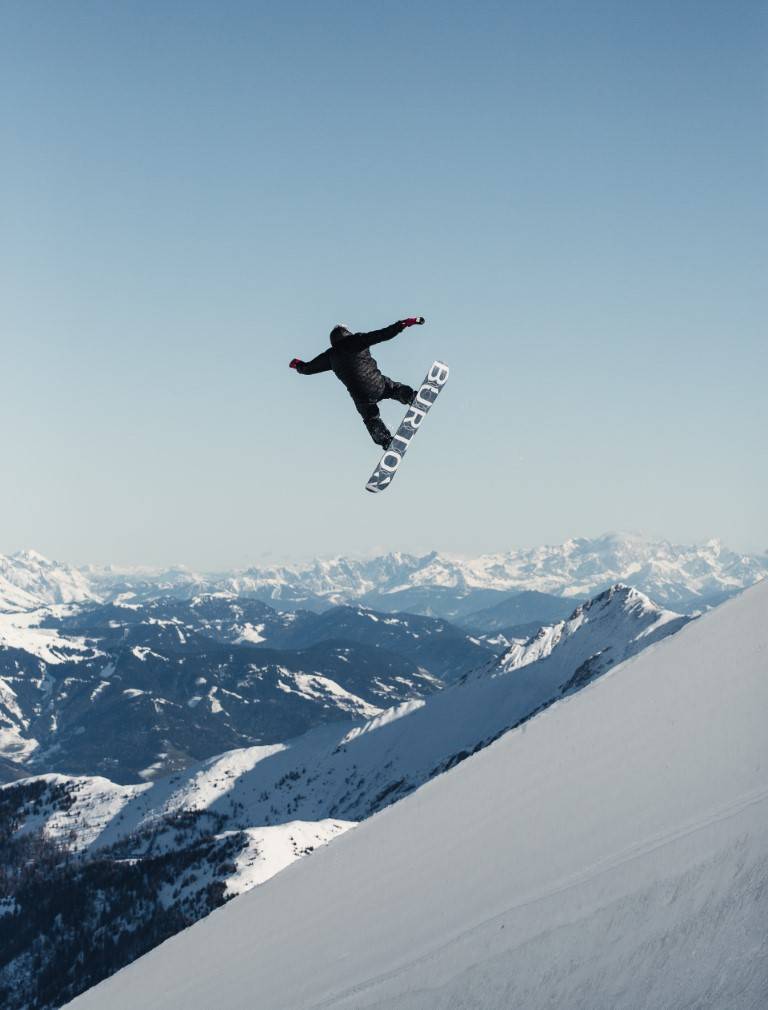 Snowboarder in mid-air against a blue sky above a snowy mountain panorama