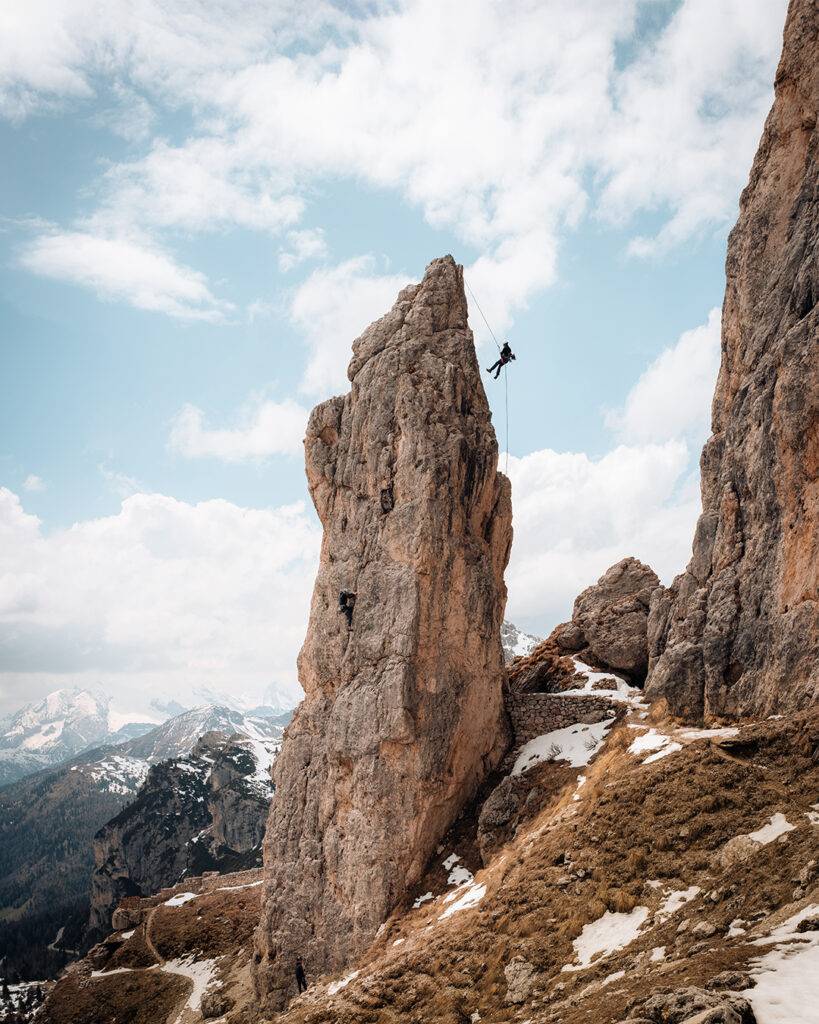 Two climbers scale a towering rock spire above snowy mountain slopes beneath a cloudy sky.