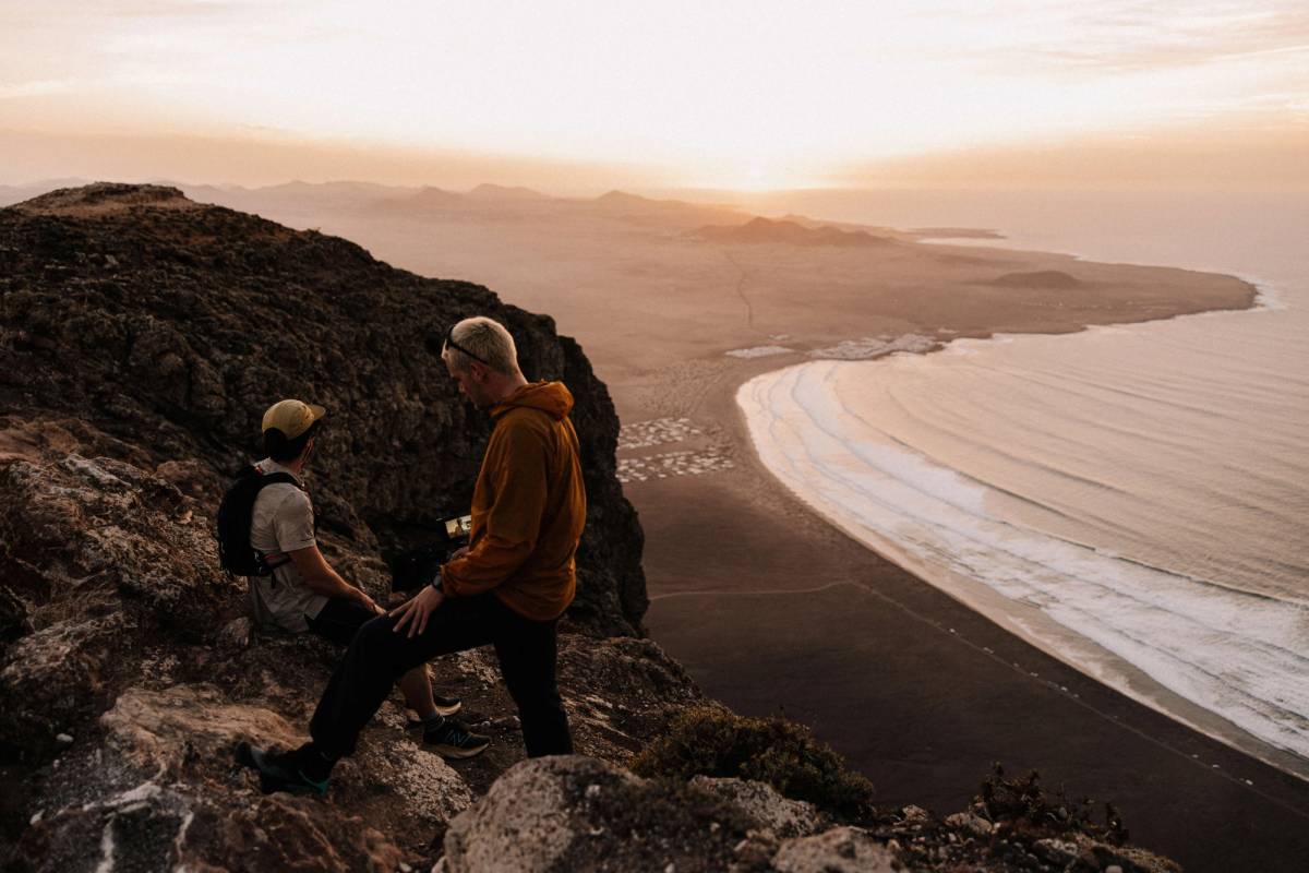 Two people with a camera on a rocky ridge above a long sandy bay in the evening light