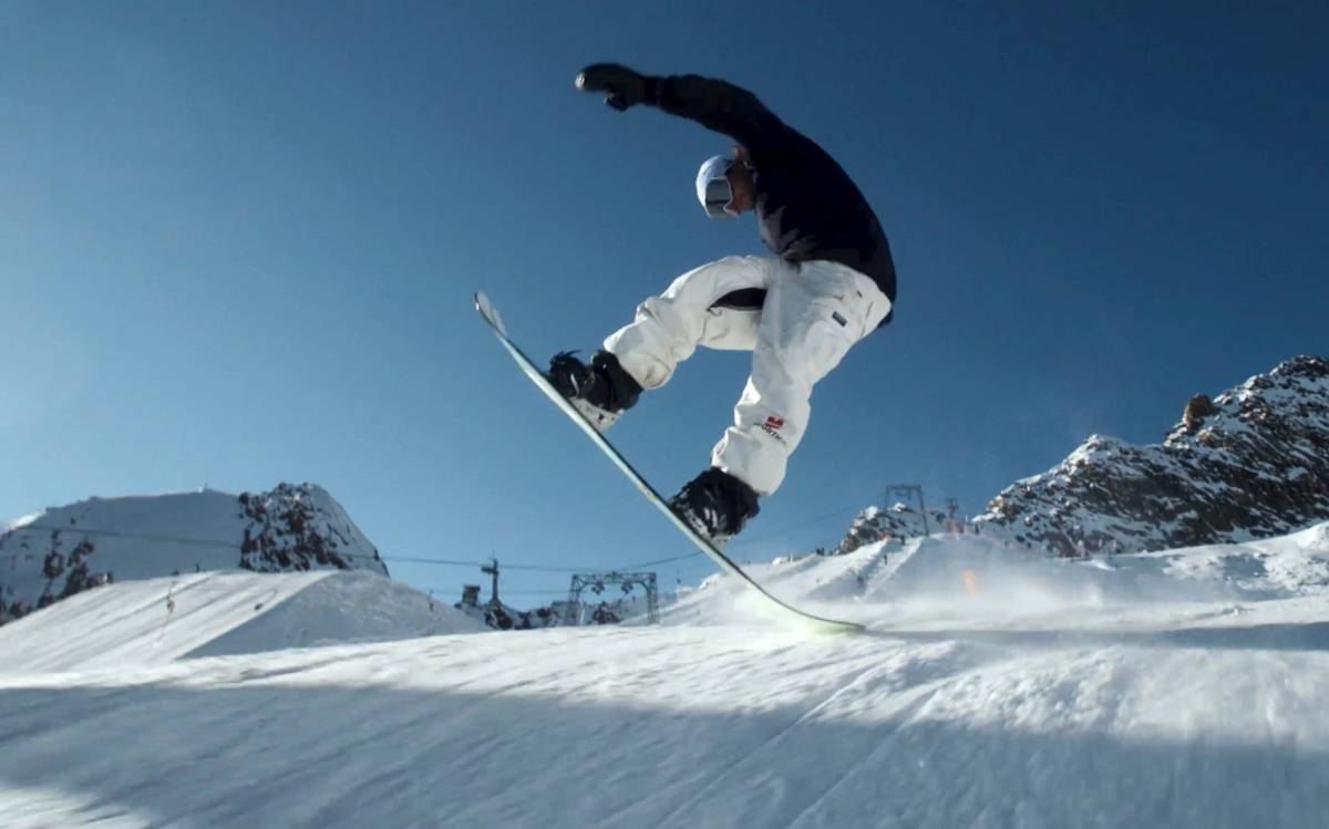 Snowboarder greift im Sprung über dem Snowpark an sein Board, dahinter blauer Himmel