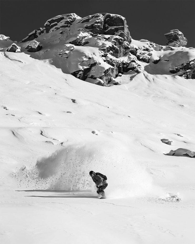 A snowboarder carves through deep powder below snow-covered rocky peaks in a black-and-white action shot.