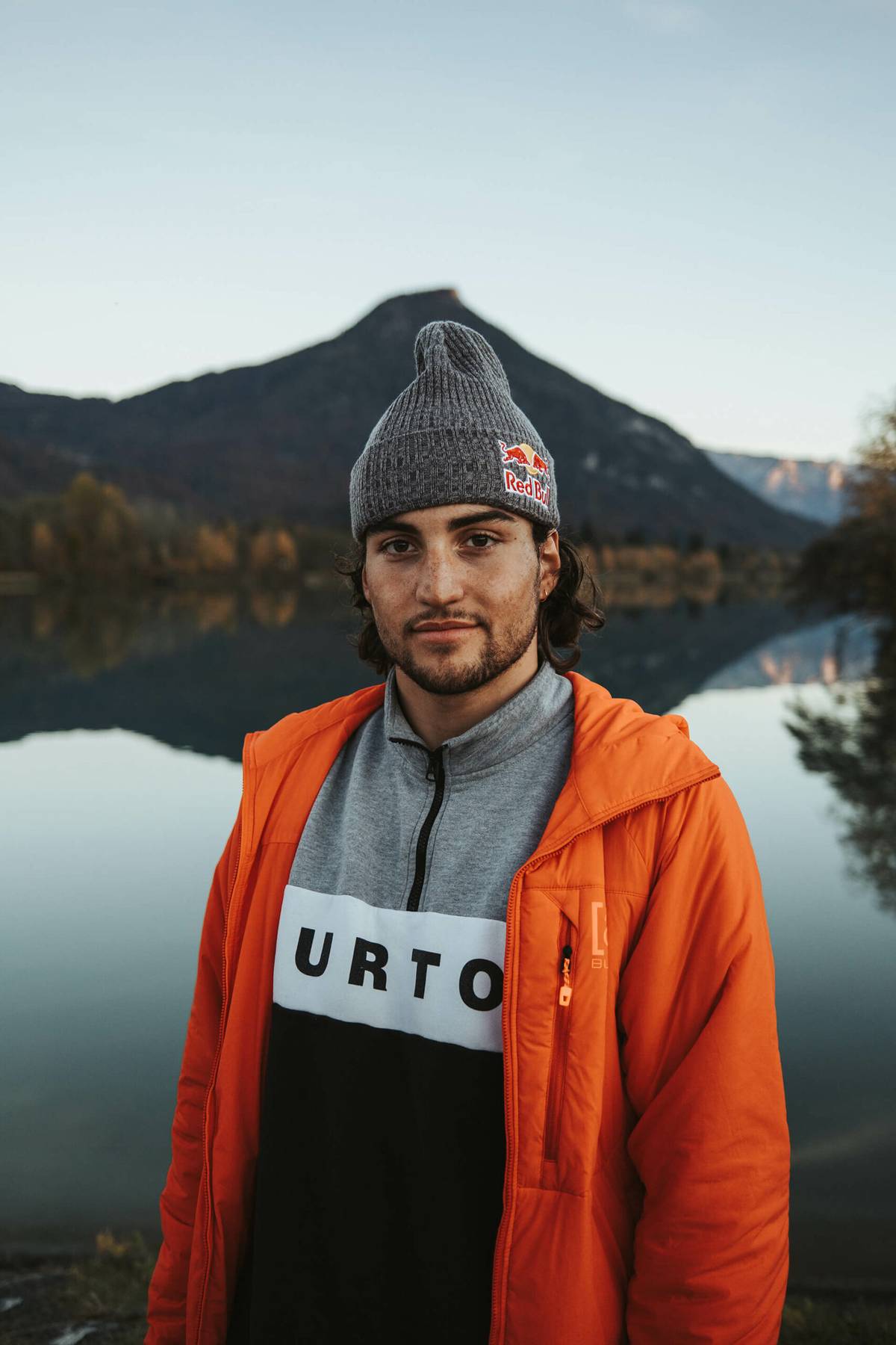 Portrait of Leon Vockensperger in a beanie and an orange jacket in front of a mountain lake