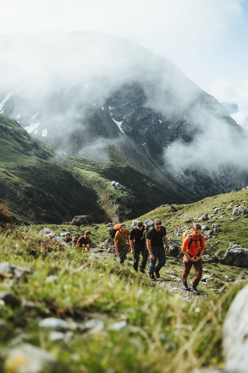 Group of hikers climbing a grassy slope in single file, cloud-covered mountains behind