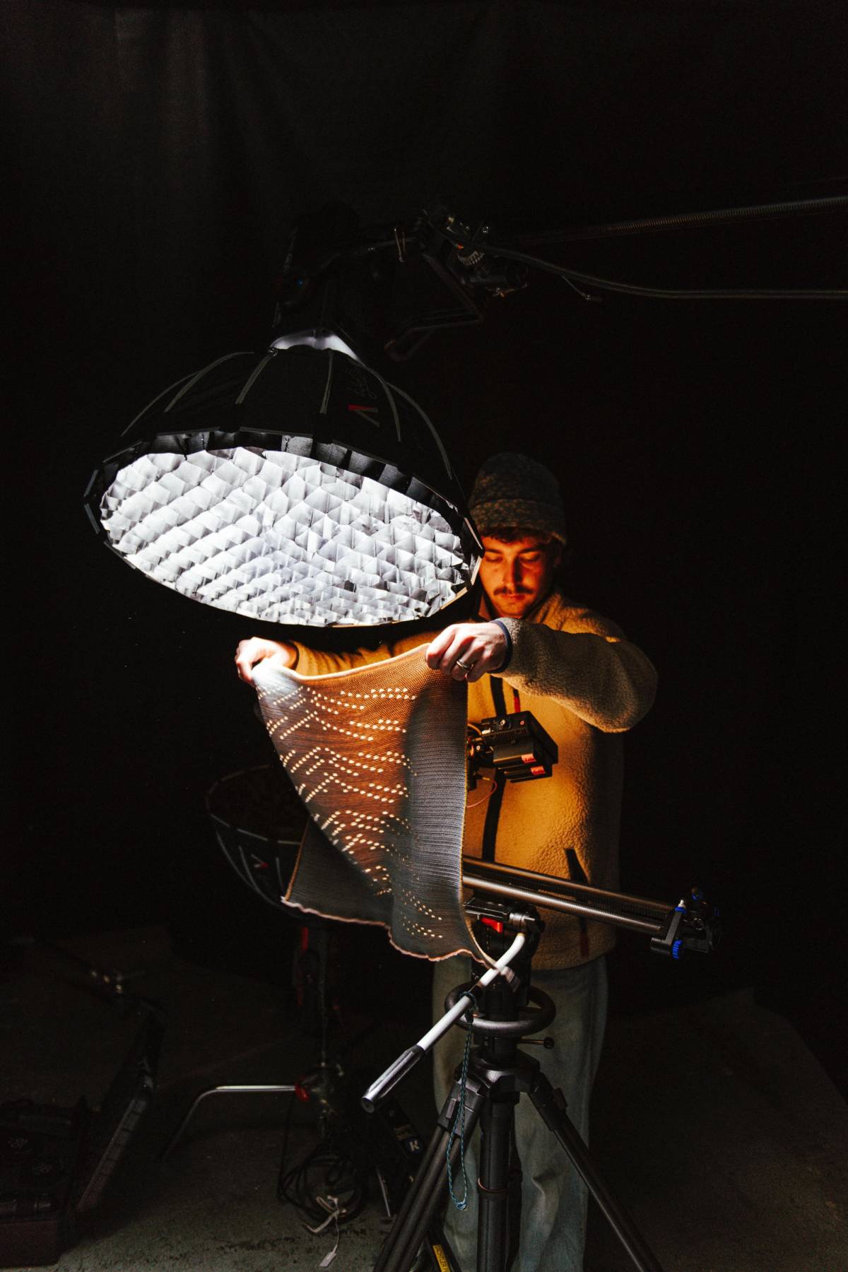 A crew member holds patterned fabric beneath a large studio light beside a camera rig in a dark production space.