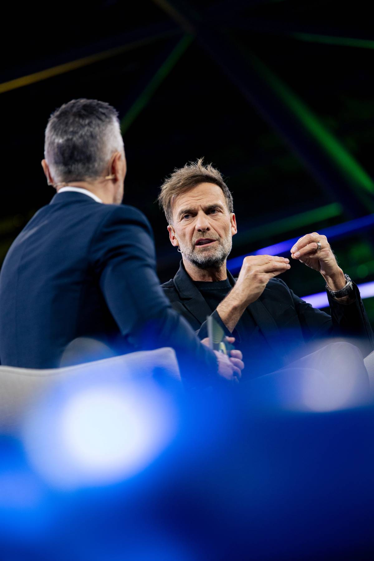 Two men wearing headsets sit on a lit stage as one gestures while speaking.