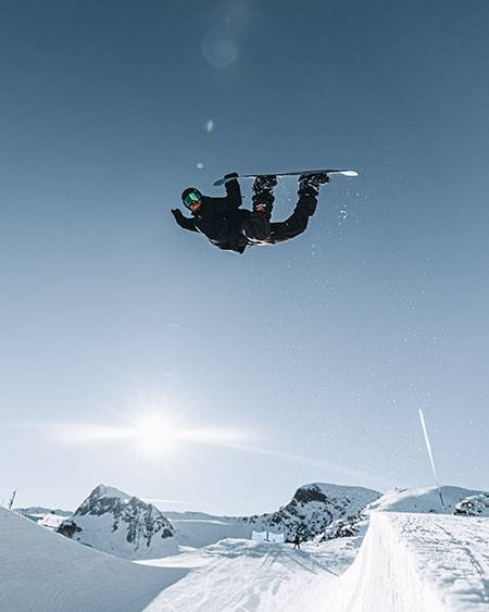 Snowboarder kopfüber in der Luft vor blauem Himmel, darunter Sonne und verschneite Berge