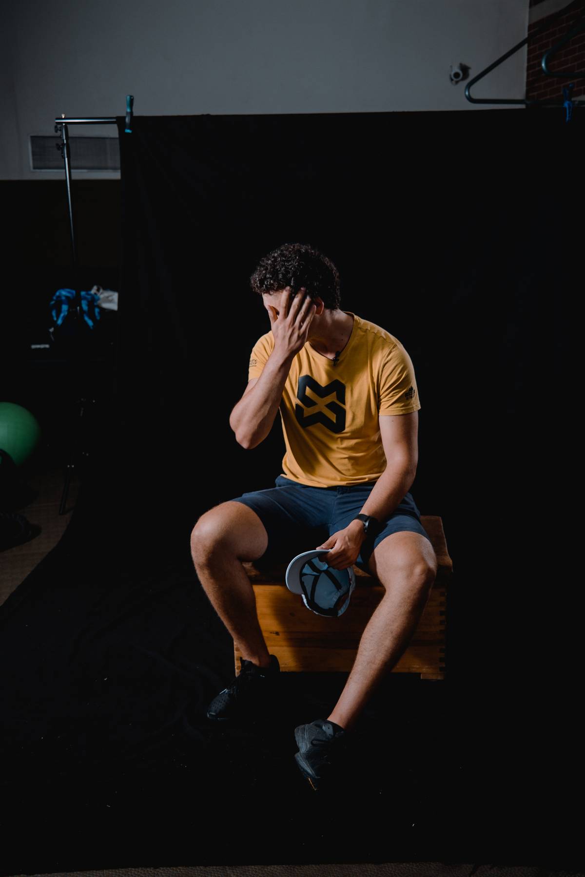 Athlete in a yellow shirt sitting on a wooden box in front of a black studio backdrop, one hand over the face