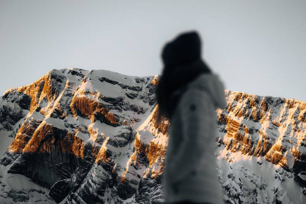 An out-of-focus person in winter clothing stands before sunlit, snow-covered mountains.