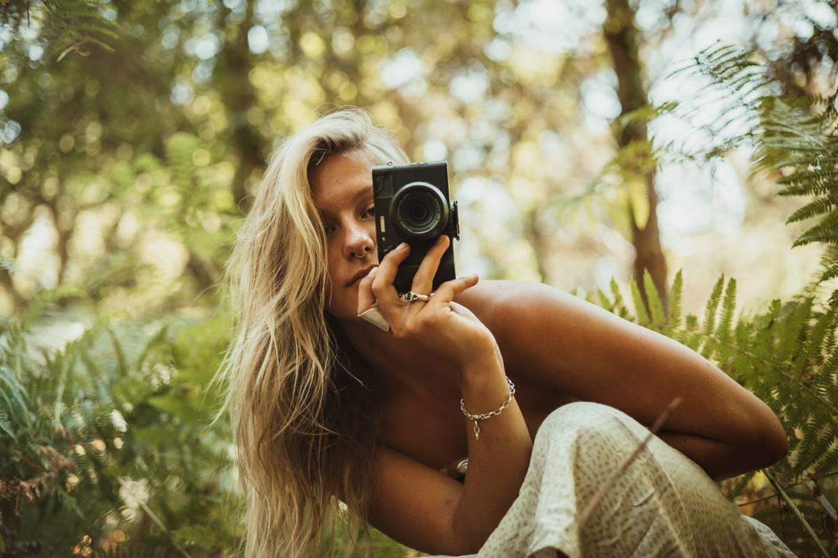 Person sitting among ferns, pointing a compact camera at the viewer