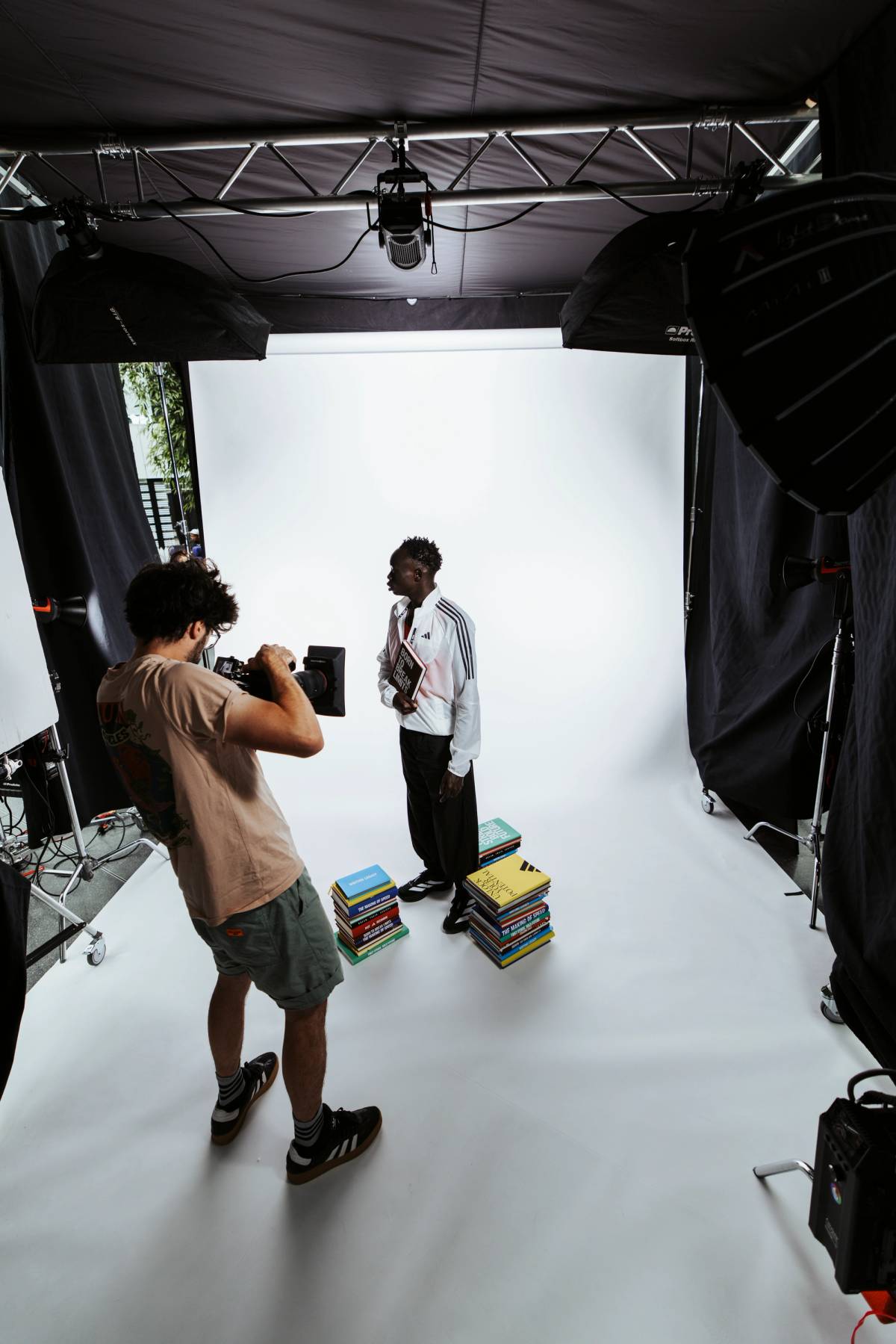 A camera operator films a man holding a book beside stacked books on a white studio set surrounded by softbox lights.