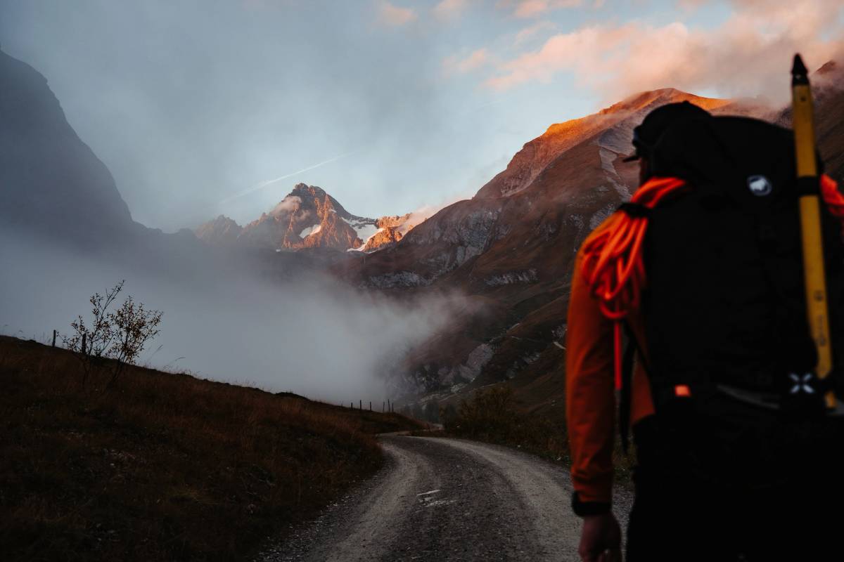 Mountaineer with a red backpack seen from behind on a gravel track, the peaks in warm light