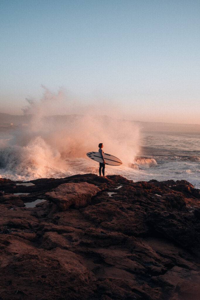 A surfer holding a surfboard stands on rocky shore as waves crash behind them in warm evening light.