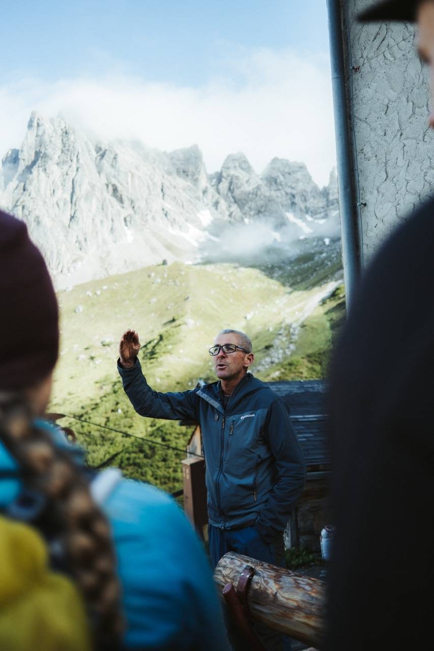 Person with glasses explaining something with a raised hand, listeners blurred in the foreground, rocky peaks behind