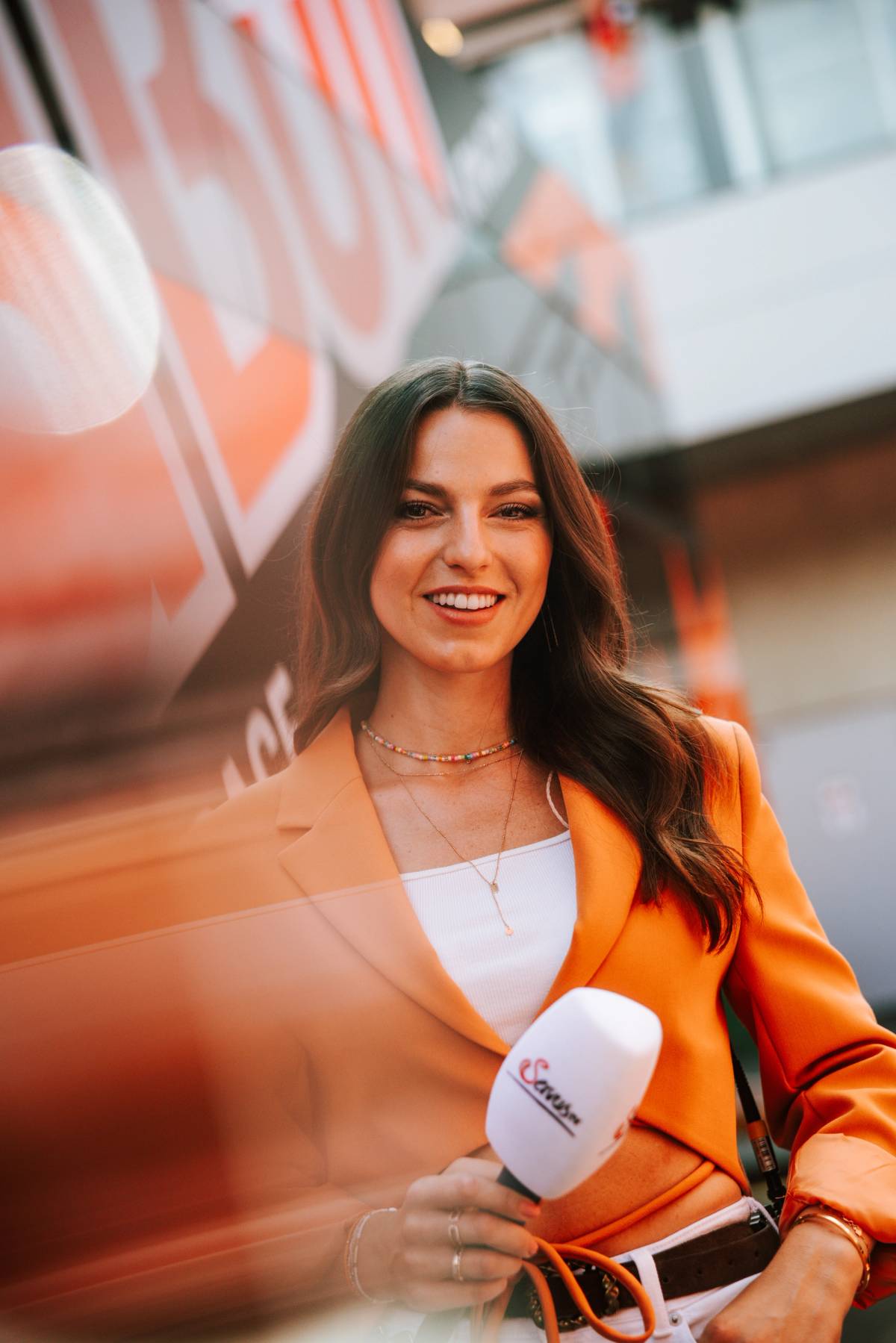 A smiling presenter in an orange blazer holds a microphone amid warm lighting and blurred production equipment.