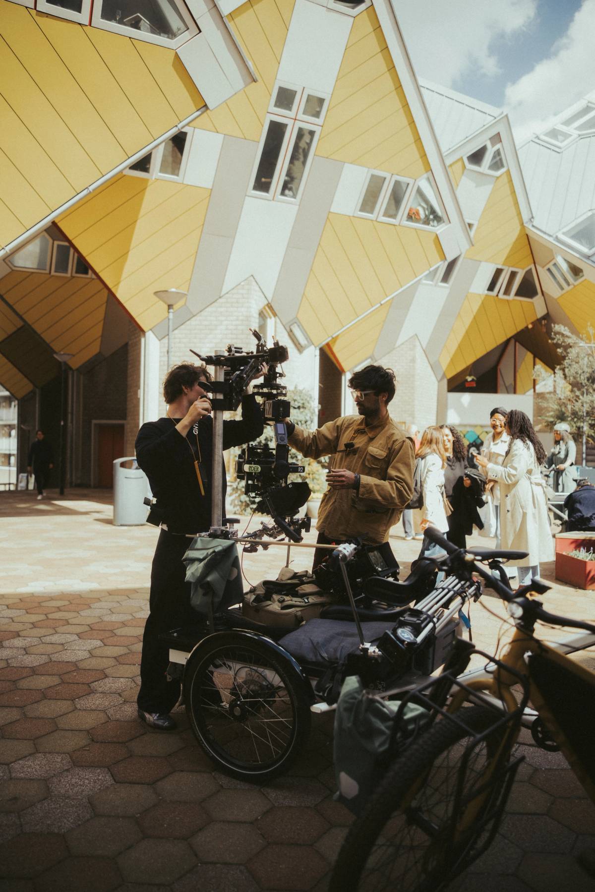 Two crew members adjust a large camera rig beside bicycles in front of angular yellow buildings.